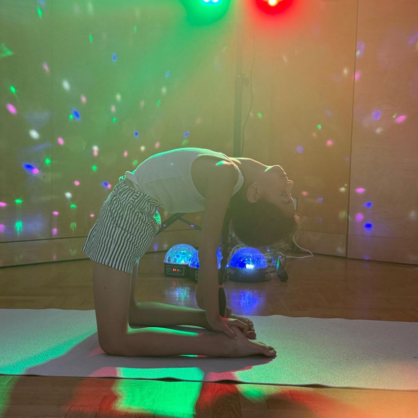 A woman is doing yoga on a mat on a wooden floor, illuminated by colorful lights. She is in a kneeling position with her head down. There are two disco balls with lights on the floor.