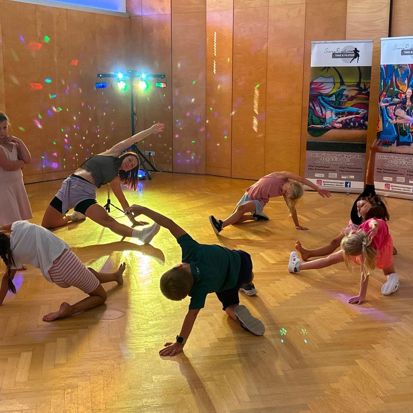 A group of children and a woman are doing yoga poses in a room with wooden floors and colorful lights.