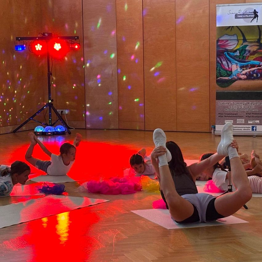 Eine Gruppe von Kindern und Erwachsenen praktiziert Yoga auf Matten auf einem Holzboden mit bunten Lichtern und einer Discokugel im Hintergrund.