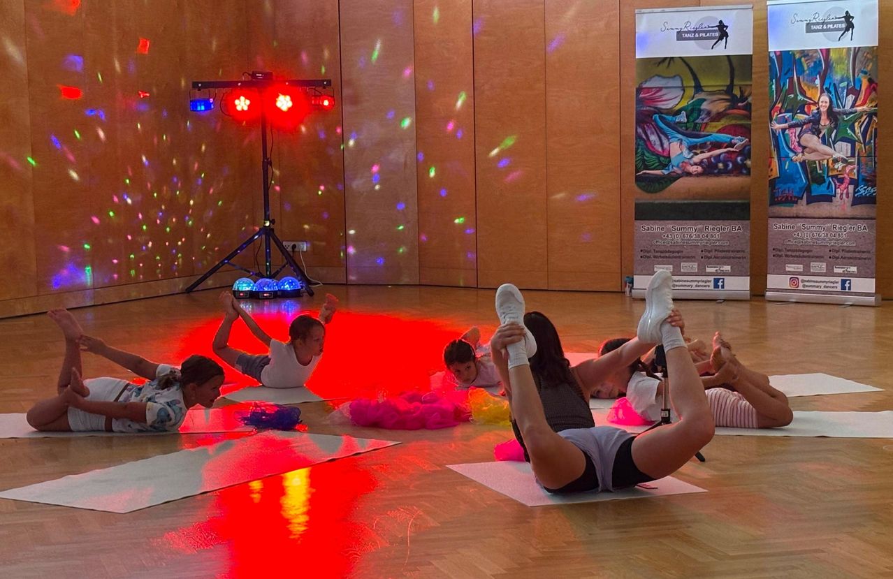 A group of children and adults are doing yoga on mats on a wooden floor with colorful lights and a disco ball in the background.