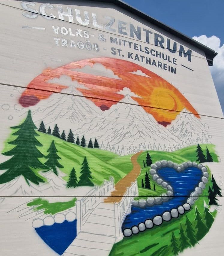 A mural on a building depicts a serene landscape with mountains, a river, and trees. The mural includes the text 'Schulzentrum Volks- und Mittelschule Tragöb - St. Katherien'.