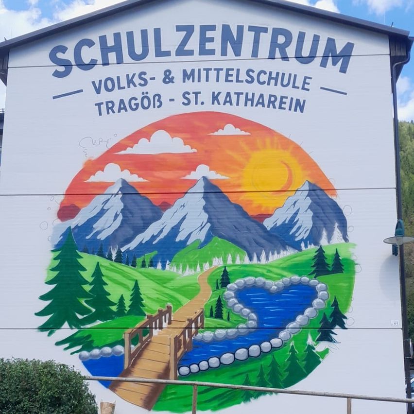 Ein Wandbild an einem Gebäude mit dem Text 'Schulzentrum Volks- & Mittelschule Tragöß - St. Kathrein'. Das Wandbild zeigt eine Berglandschaft mit einem Fluss und einer Brücke.
