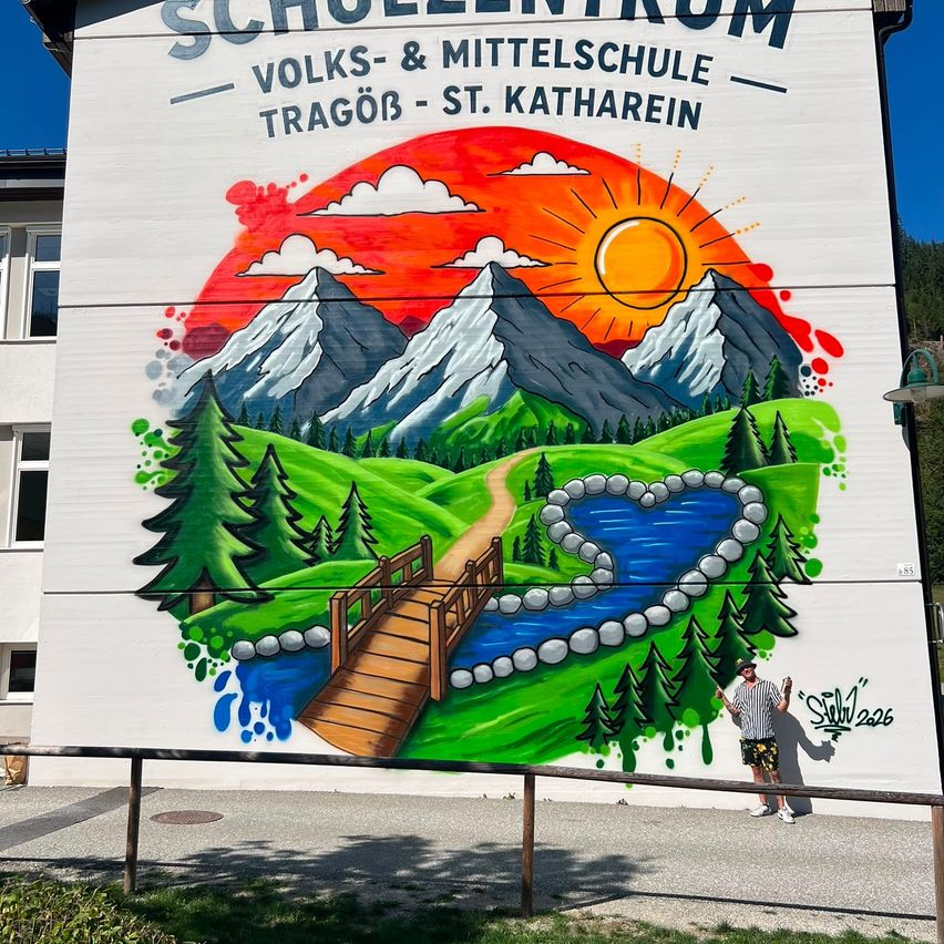 Ein Wandbild auf einem Gebäude zeigt eine friedliche Landschaft mit Bergen, einem Fluss und einer Brücke unter einem untergehenden Sonnen. Eine Person steht in der Nähe und posiert für ein Foto.
