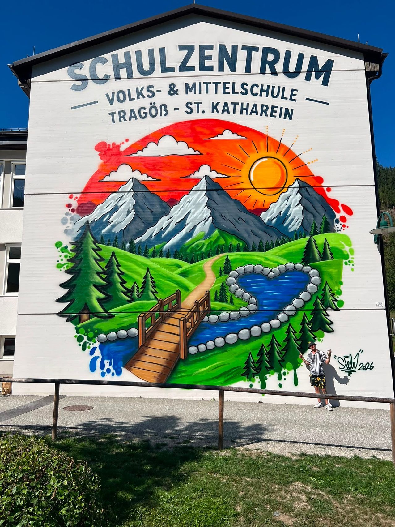 A mural on a building depicts a serene landscape with mountains, a river, and a bridge under a setting sun. A person stands nearby, posing for a photo.