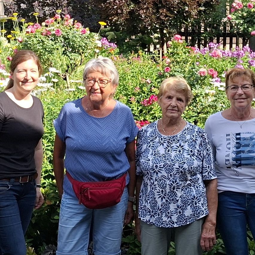 Four women stand in a garden, smiling for a photo. The woman on the left wears a black shirt and jeans. The woman next to her wears a blue blouse and a red fanny pack. The third woman wears a floral-printed top and the fourth wears a white shirt with a graphic design.
