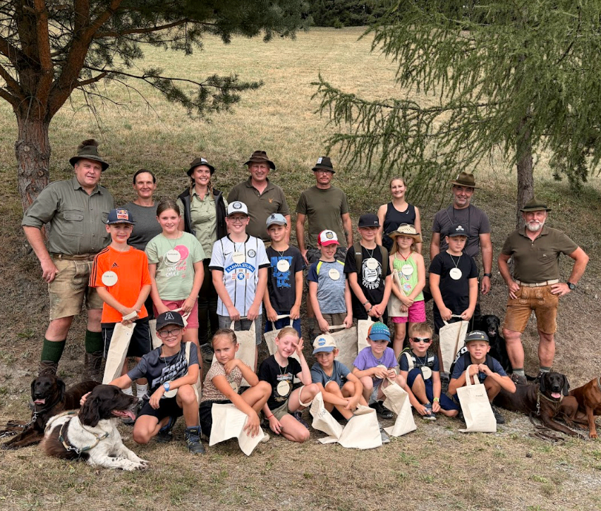 Eine Gruppe von Erwachsenen und Kindern, alle mit Hüten, stehen auf einem Feld mit Bäumen im Hintergrund. Sie halten Taschen und einige tragen Medaillen. Zwei Hunde sitzen vor ihnen.