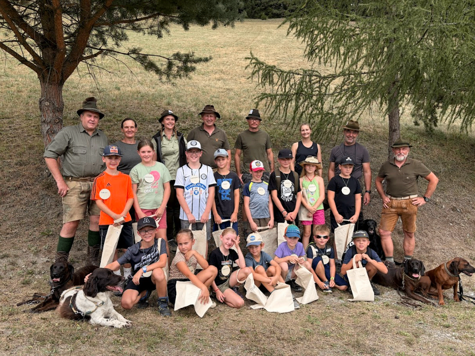 A group of adults and children, all wearing hats, stand in a field with trees in the background. They are holding bags and some are wearing medals. Two dogs are sitting in front of them.