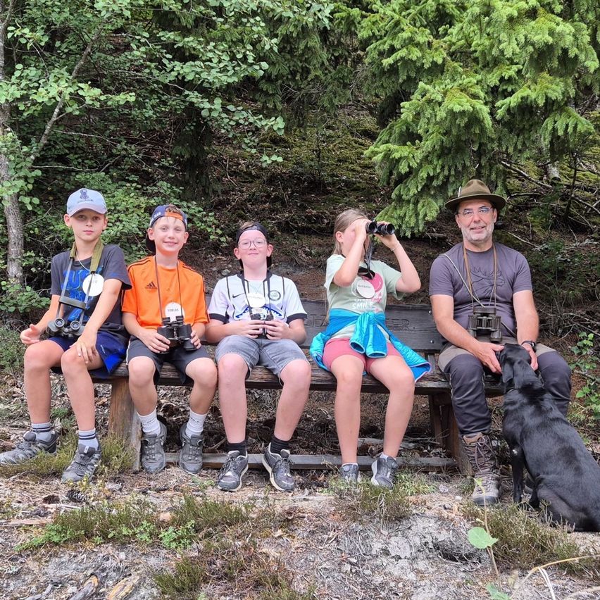 Eine Familie mit fünf Mitgliedern, darunter ein Mann, eine Frau und drei Kinder, sitzt auf einer Bank im Wald und schaut alle mit Ferngläsern in die Ferne.