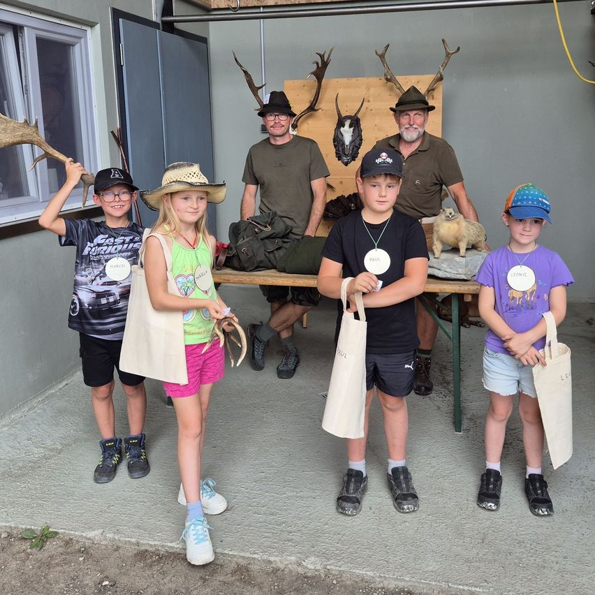 A group of children and two men stand in front of a table with antlers and a marmot. The children hold bags and wear name tags.