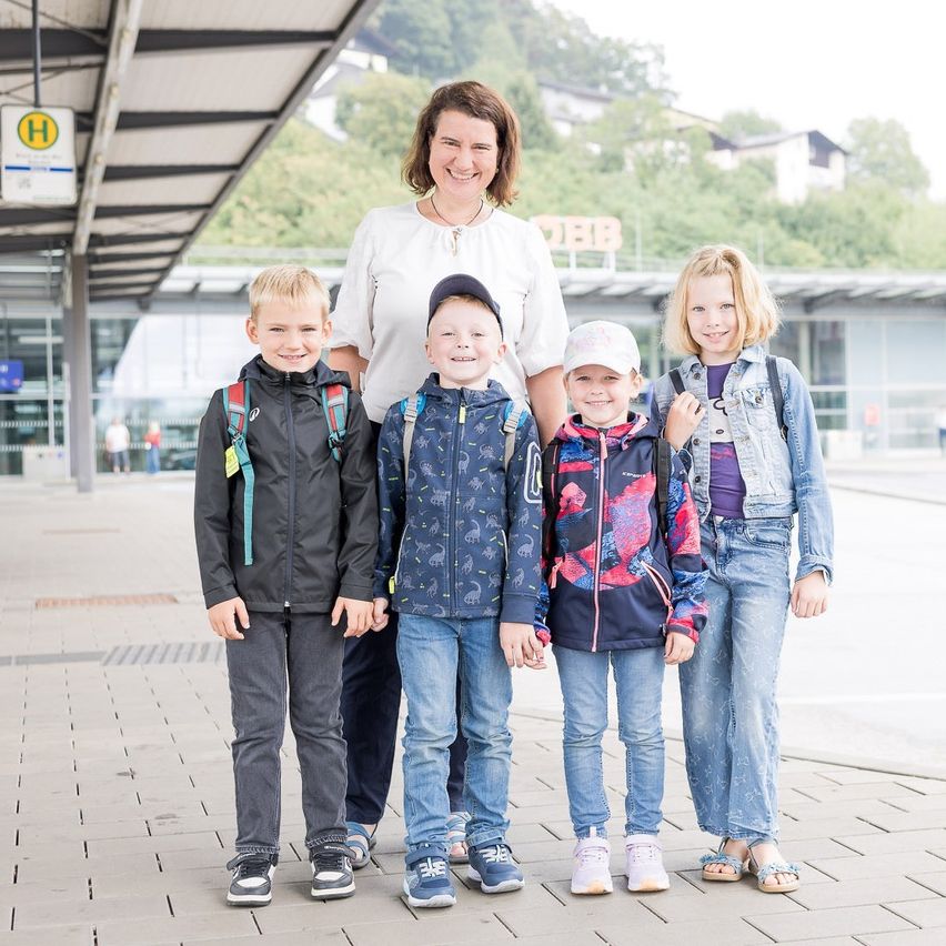 Eine Frau und vier Kinder stehen lächelnd auf einem Ziegelstein-Gehweg und posieren für ein Foto. Die Kinder tragen Rucksäcke und Turnschuhe. Hinter ihnen ist ein Gebäude mit einem Schild zu sehen.