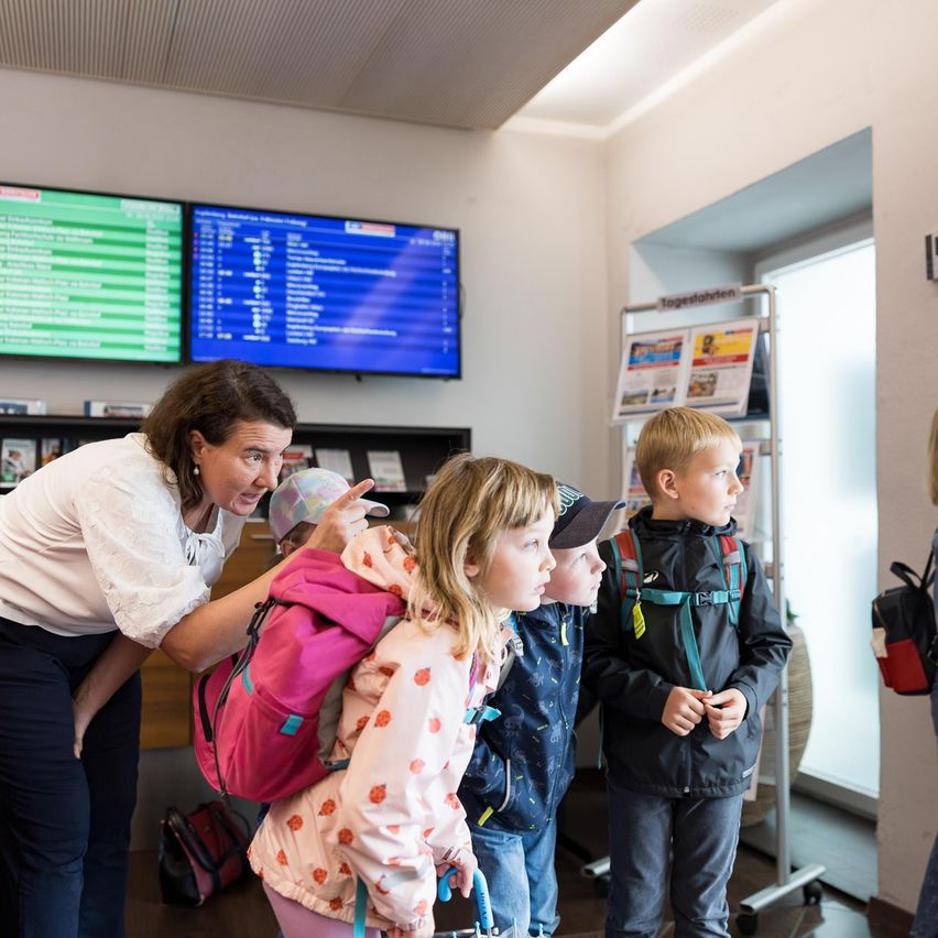 Eine Frau mit einem Rucksack hilft einem Mädchen mit einer rosa gepunkteten Jacke und einem Jungen mit Hut in einer Wartehalle. Sie stehen vor einem digitalen Board und einer Glastür.