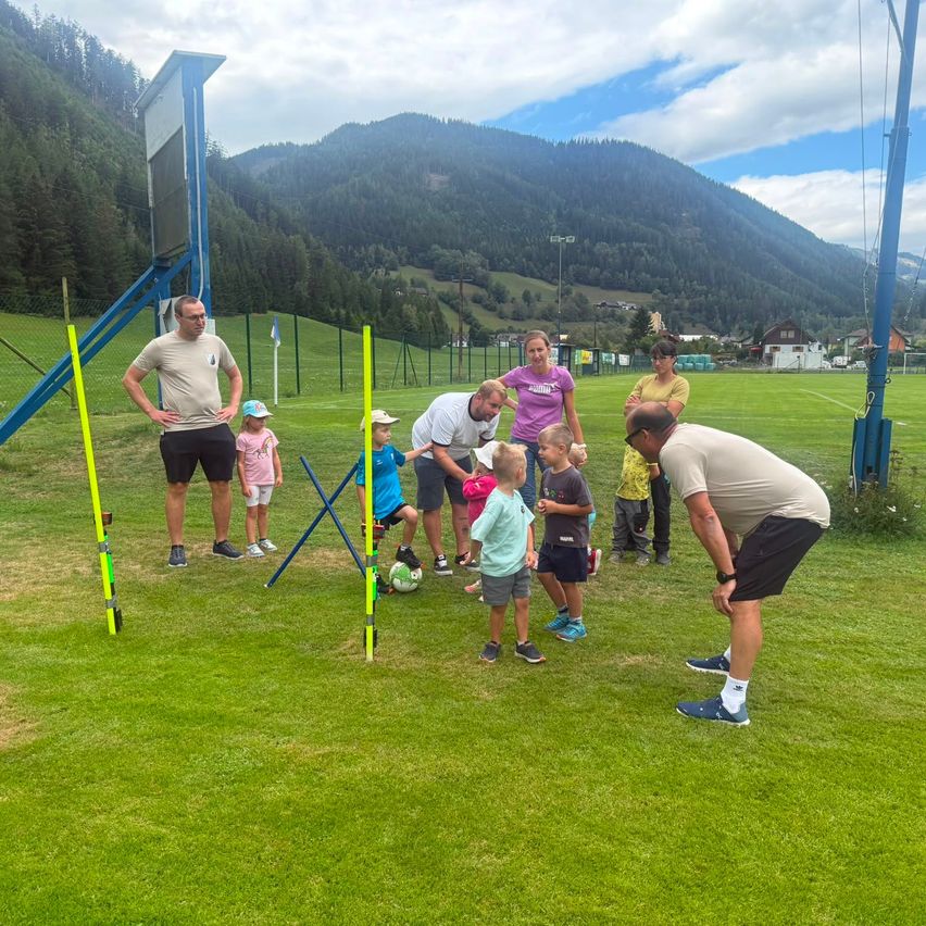 Eine Gruppe von Erwachsenen und Kindern spielt Fußball auf einem Feld mit einem Berg im Hintergrund.