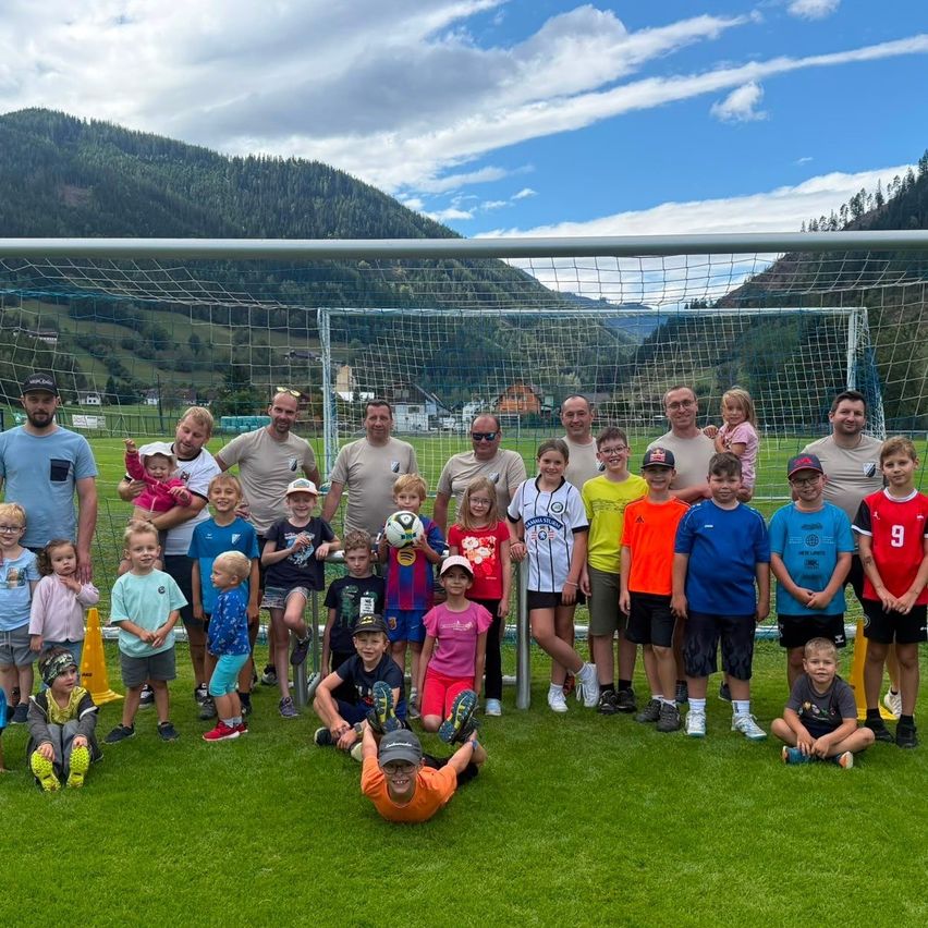 Eine Gruppe von Kindern und Erwachsenen posiert für ein Foto vor einem Fußballtor mit einem Gebirgshintergrund.
