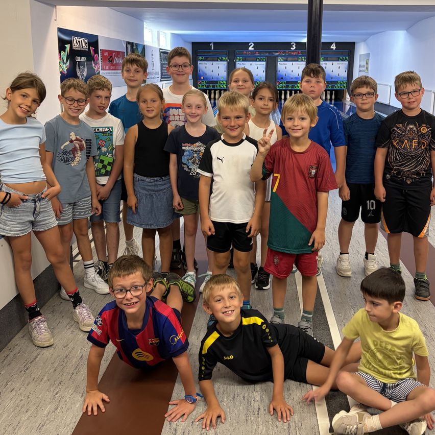Eine Gruppe von Kindern in einer Bowlingbahn, einige stehend und einige sitzend, lächeln für ein Foto.
