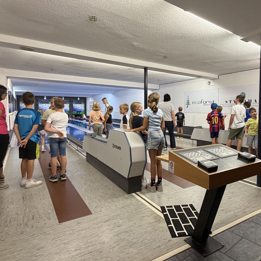Eine Gruppe von Kindern und Erwachsenen versammelt sich um eine Bowlingbahn und beobachtet ein Spiel. Die Bowlingbahn ist gut beleuchtet und hat ein modernes Design.