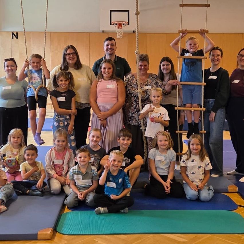 Eine Gruppe von Kindern und Erwachsenen posiert für ein Foto in einer Turnhalle. Einige Kinder sitzen auf Yogamatten, während andere stehen oder eine Leiter erklimmen.