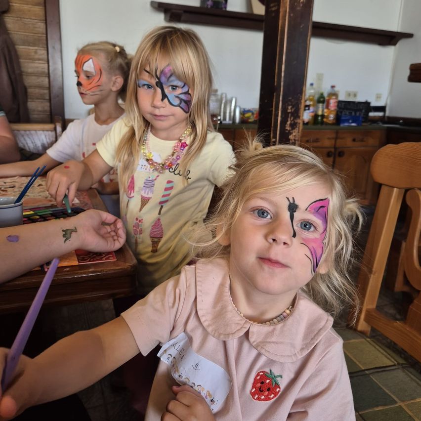 Drei junge Mädchen mit Gesichtsbemalung sitzen an einem Tisch, eine mit Schmetterlings-Gesichtsbemalung und die andere mit Tiger-Gesichtsbemalung.