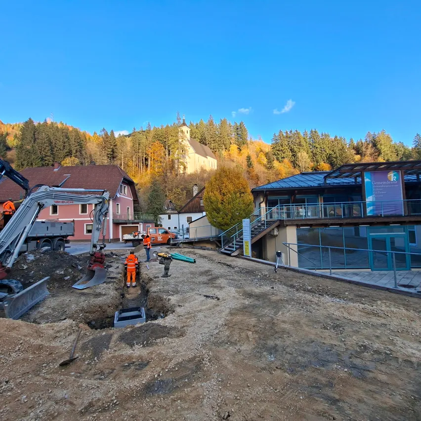 Eine Baustelle mit schwerem Gerät und Arbeitern in orangefarbenen Uniformen. Die Baustelle ist von Häusern und einer Kirche umgeben, mit einem klaren blauen Himmel und Bäumen im Hintergrund.