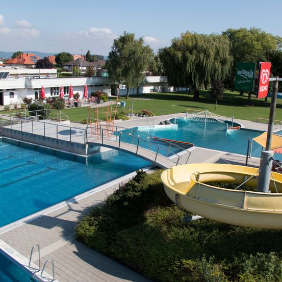 Luftaufnahme eines Schwimmbeckens mit einer gelben Wasserrutsche. Es ist von einem Zaun und einer Rasenfläche umgeben. Im Hintergrund befinden sich Gebäude und Bäume.