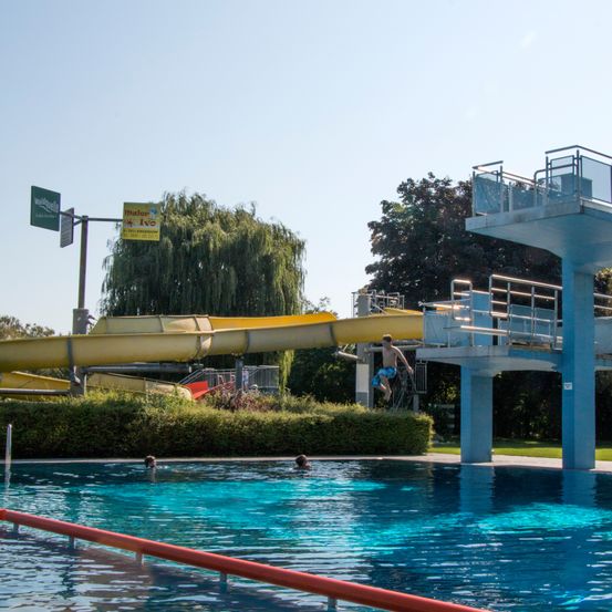Eine große gelbe Wasserrutsche und eine Sprungplattform neben einem Freischwimmbecken mit Schwimmenden.