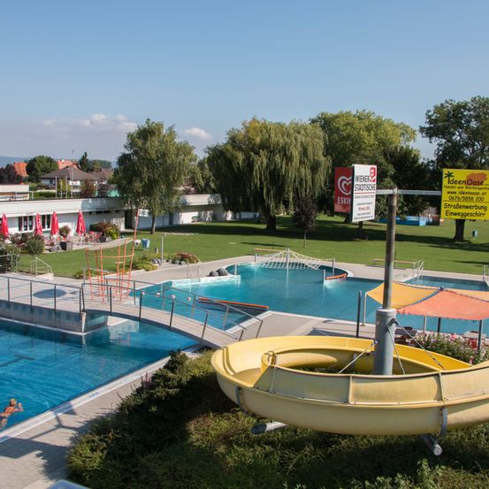 Eine Vogelperspektive auf einen großen Außenpoolkomplex zeigt eine Wasserrutsche und eine Sprungbrett. Schilder weisen auf 'Wiener Stadtische' und 'IdeenOase' hin. Der Bereich ist von viel Grün umgeben und der Himmel ist klar.