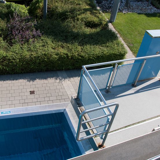 Ein Luftbild eines Pools mit einem blauen Metallgeländer und einem gefliesten Gehweg. Der Pool ist von einer Hecke und einem Grasbereich umgeben.