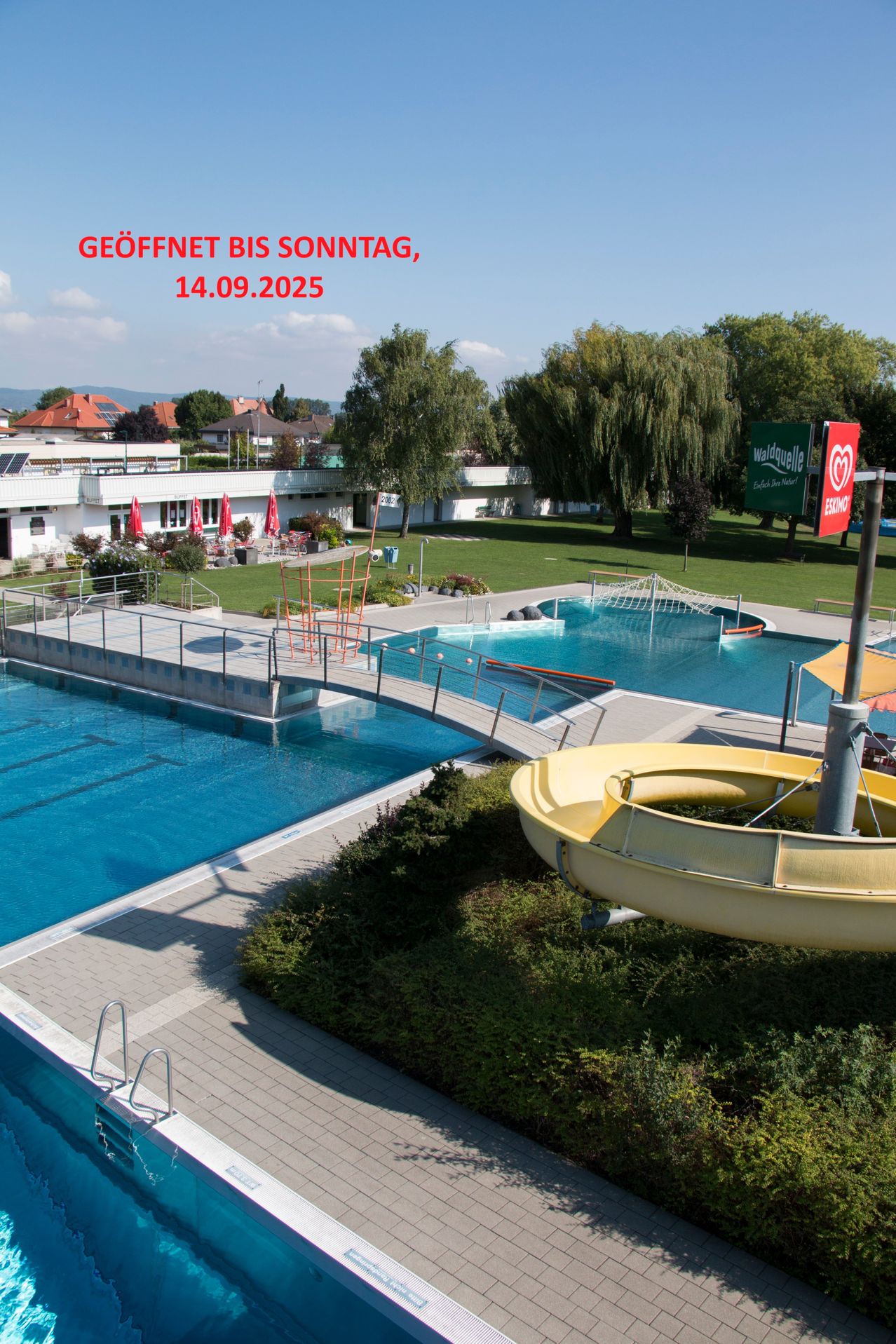 Ein Blick von oben auf einen großen Swimmingpool mit einer gelben Wasserrutsche und einer Brücke. Es gibt ein Schild mit Text auf Deutsch und einem Datum.