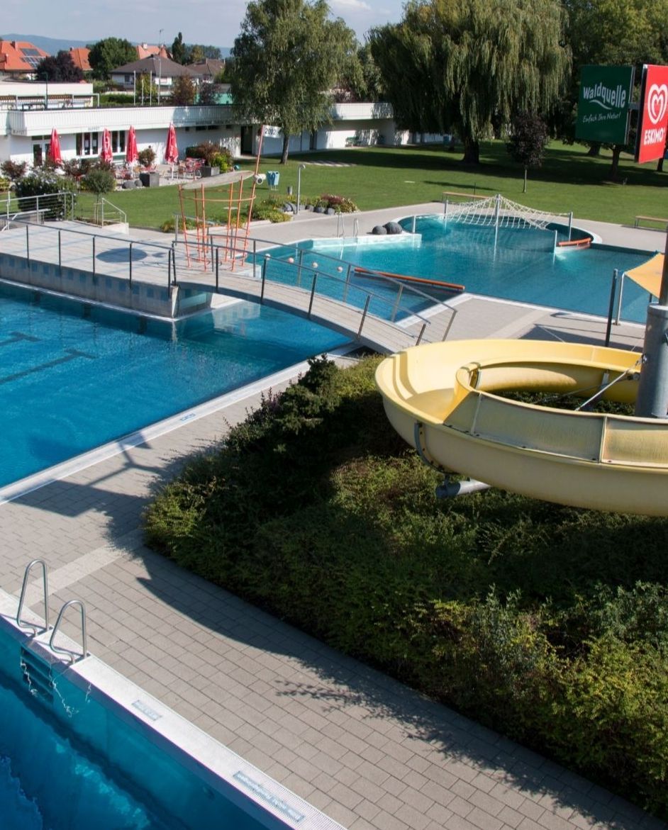 Ein Freibadbereich mit zwei Schwimmbecken, einer gelben Wasserrutsche und einem Gebäude mit roten Sonnenschirmen.