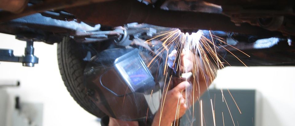 Eine Person schweißt die Unterseite eines Autos, trägt eine Schweißmaske und Handschuhe. Funken fliegen umher.