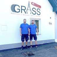 Zwei Männer in blauen Uniformen stehen vor einem weißen Gebäude mit dem Wort 'Grass' darauf.