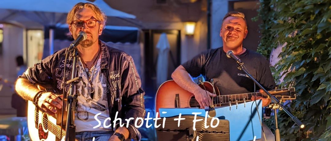 Zwei Männer spielen im Freien, einer spielt Gitarre und singt, der andere spielt eine Keyboard. Im Hintergrund befindet sich ein Gebäude mit einer leuchtenden Lampe. Der Text lautet 'Schrotti + Flo Ostpop unplugged'.