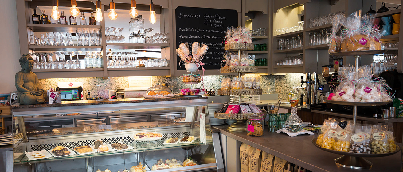 Eine Bäckerei mit einer Vitrine voller verschiedener Gebäcke. Ein Kreidetafel-Menü hängt an der Wand. Über dem Tresen sind mehrere Teller und Gläser angeordnet.