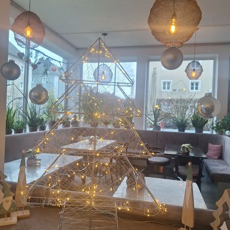 Ein Essbereich mit einem beleuchteten Weihnachtsbaum in der Mitte, umgeben von Tischen und Stühlen. Im Hintergrund befinden sich Topfpflanzen und Hängelampen. Das Bild zeigt auch ein Gebäude mit Fenstern im Freien.