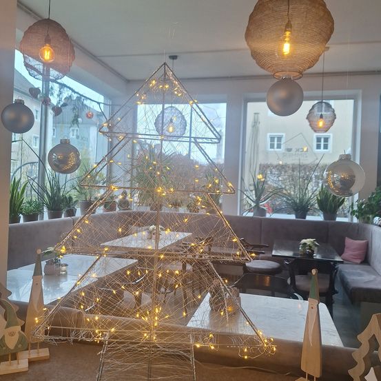 Ein Essbereich mit einem beleuchteten Weihnachtsbaum in der Mitte, umgeben von Tischen und Stühlen. Im Hintergrund befinden sich Topfpflanzen und Hängelampen. Das Bild zeigt auch ein Gebäude mit Fenstern im Freien.