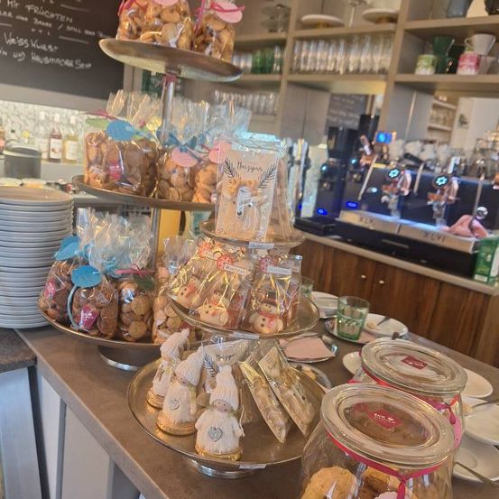 Eine Ausstellung verschiedener verpackter Süßigkeiten und Kekse in einem Geschäft. Links ein Stapel Teller. Dahinter ein Regal mit Flaschen, einer Kaffeemaschine und einer Tafel.