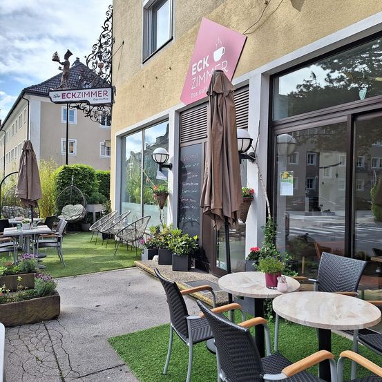 Ein Cafébereich im Freien mit Tischen und Stühlen, geschmückt mit Pflanzen und Sonnenschirmen. Das Gebäude hat ein Schild mit der Aufschrift 'ECK ZIMMER'. Die Atmosphäre ist gemütlich mit grünem Gras und einem einladenden Eingang.