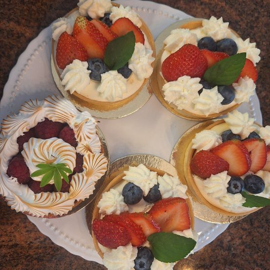 Fünf Törtchen auf einem weißen Teller. Jedes Törtchen hat Schlagsahne und ist mit Erdbeeren, Himbeeren oder Blaubeeren garniert.