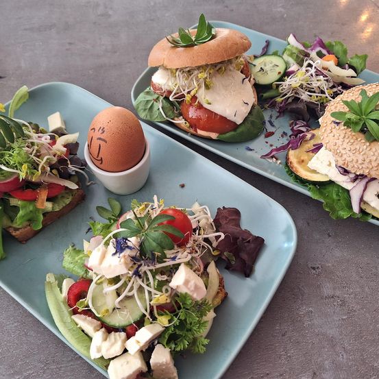 Drei blaue Teller enthalten Sandwiches und Salate. Ein Sandwich hat ein spielerisches Ei mit einem Gesicht darauf.