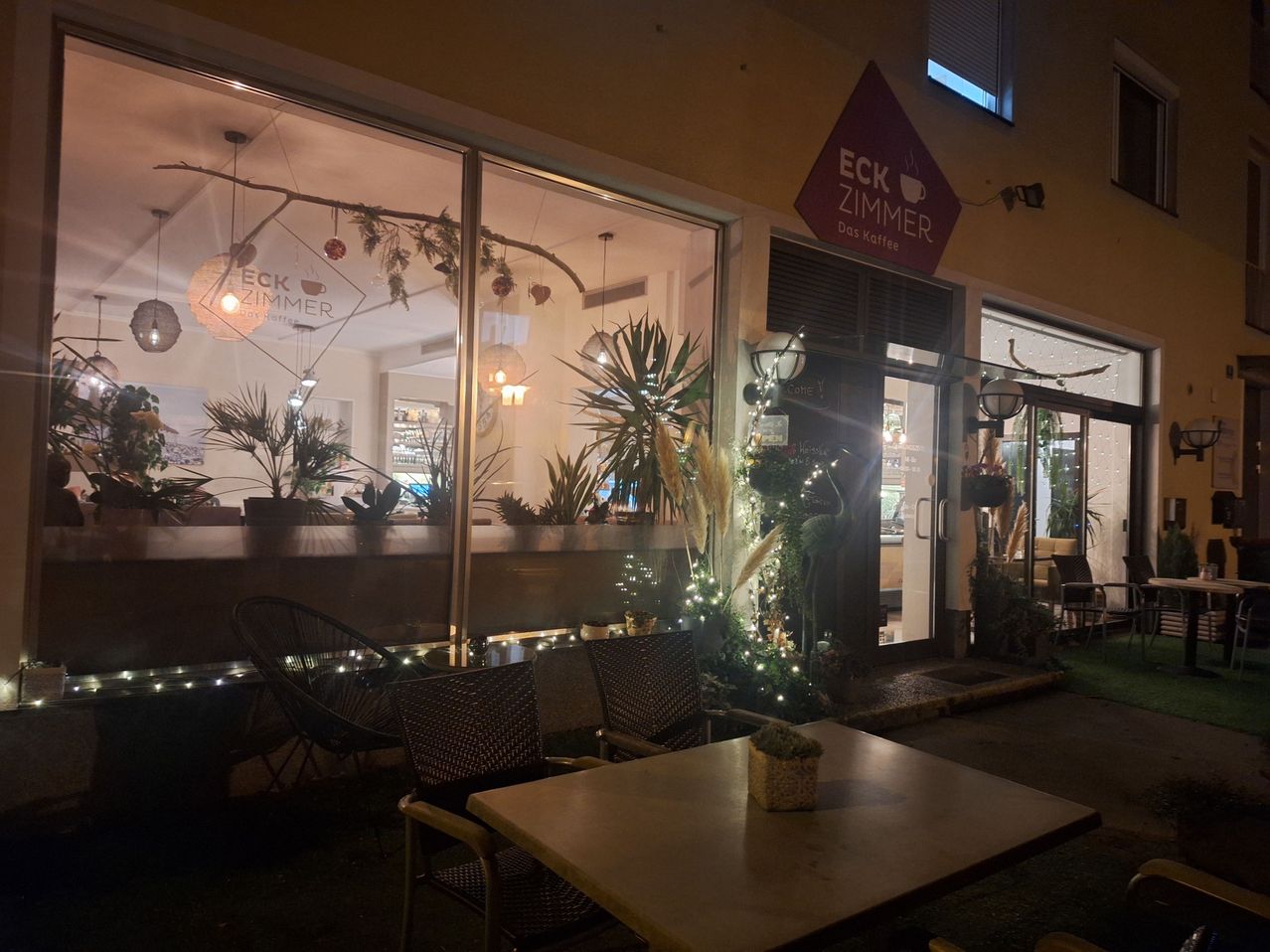 Ein Café-Eingang mit einem Schild 'ECK ZIMMER' ist bei Nacht beleuchtet. Der Außenbereich hat Korbstühle um einen Tisch, mit Pflanzen und Lichtern.