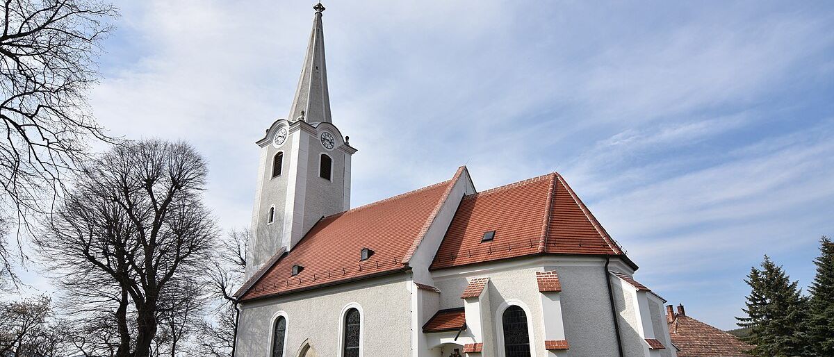 Eine weiße Kirche mit einem roten Dach und einem hohen Turm. Eine Uhr ist auf dem Turm sichtbar. Eine Statue steht vor dem Eingang.