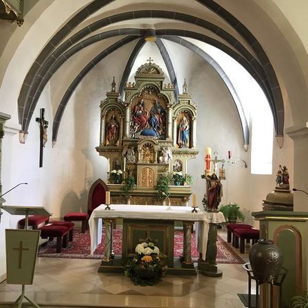 Eine Kapelle mit einem Altar, Kerzen, Blumen und religiösen Statuen. Der Altar hat einen goldenen Rahmen und ist mit religiösen Ikonen verziert. Es gibt zwei rote Stühle und einen roten Teppich auf dem Boden.