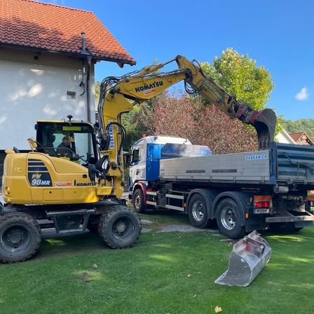 Ein gelber Komatsu-Radlader steht vor einem Haus. Ein blauer Lkw mit der Aufschrift Duskanić ist dahinter geparkt. Eine Person sitzt im Radlader.