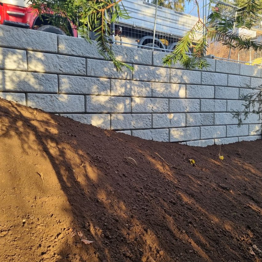 Eine Baustelle mit einem großen Haufen Erde vor einer Ziegelmauer und einem roten Fahrzeug, das an der Seite geparkt ist.