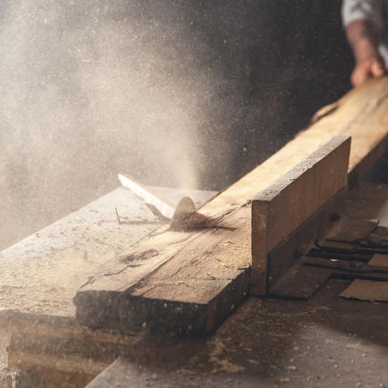 Ein Zimmermann benutzt eine Kreissäge, um ein Holzbrett zu schneiden, wobei Sägemehl in der Luft fliegt.