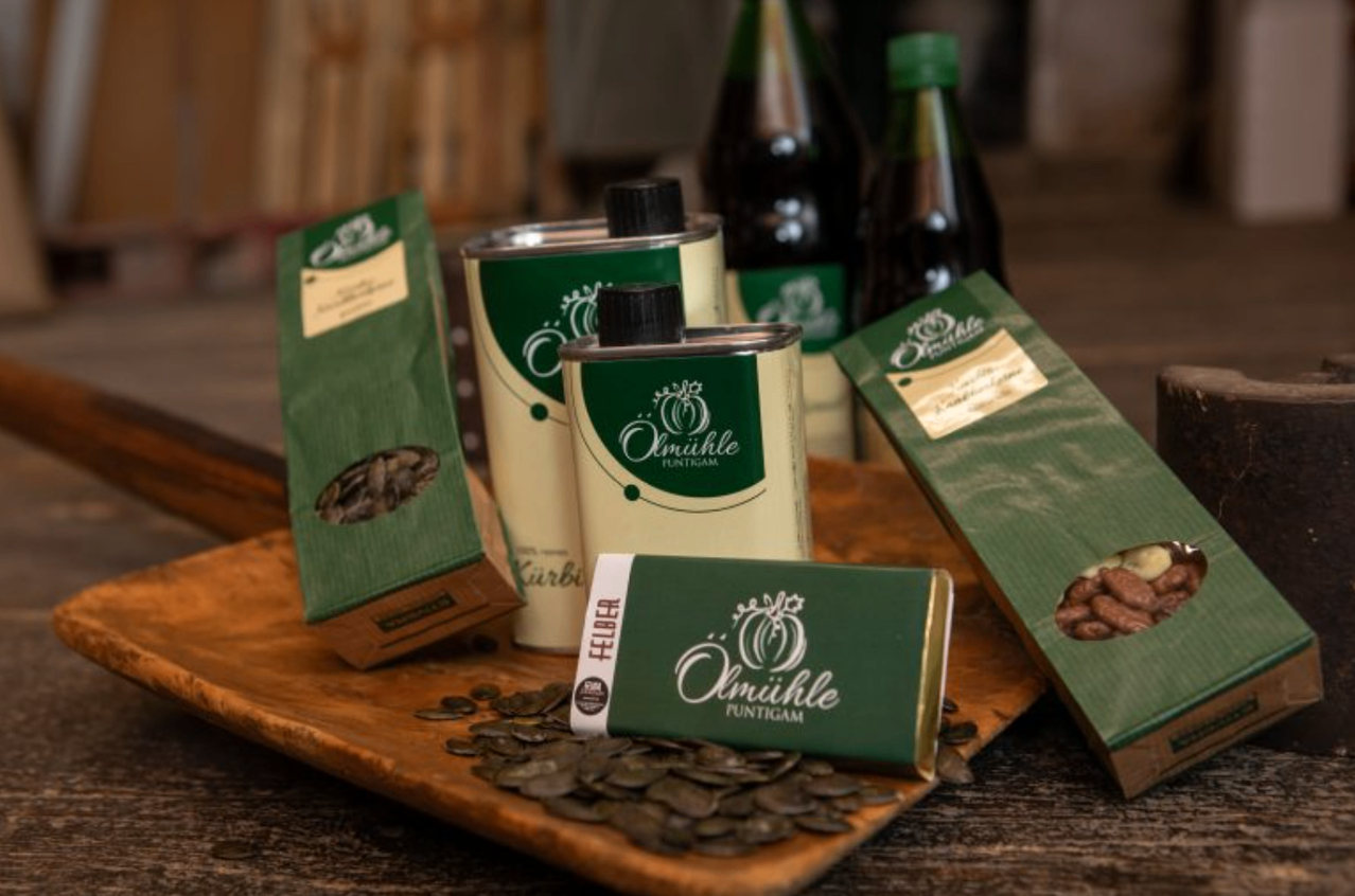 Ein Holztablett zeigt verschiedene Produkte, darunter eine grüne Dose, eine grüne Tafel und eine grüne Schachtel, alle mit dem Label 'Olmihle'. Dahinter befinden sich zwei Flaschen mit grünen Kappen.