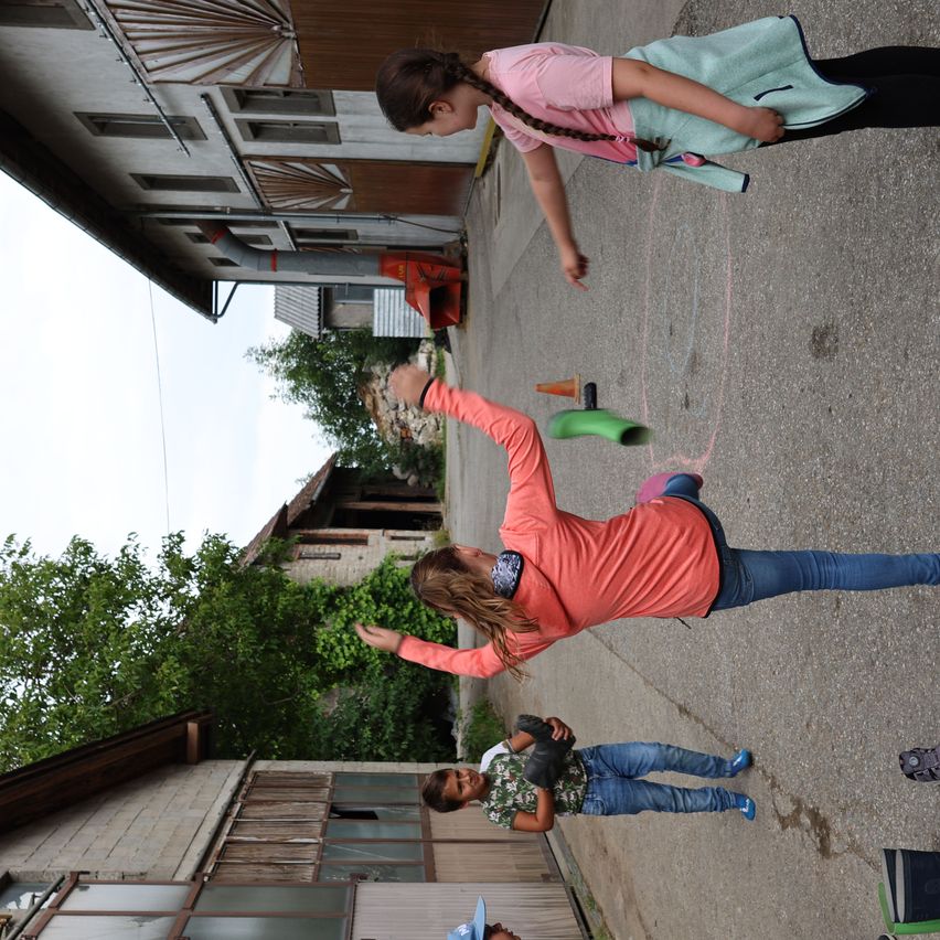 Drei Kinder spielen auf einer Straße, eines hält einen Kreidestift und malt. Die anderen beobachten. Gebäude mit Fenstern säumen die Straße, und ein Baum ist in der Nähe.
