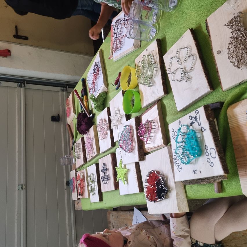 Eine Draufsicht auf einen mit grünem Stoff bedeckten Tisch, der eine Vielzahl von Holzblöcken mit aufwendigen Fadenkunstdesigns zeigt. Verschiedene Farben von Schnüren, Werkzeugen und Bechern sind auf dem Tisch verstreut. Eine Person sitzt in der Nähe und arbeitet an einem Stück.