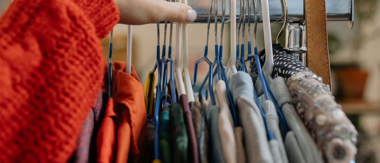 Eine Person in einem roten Pullover hält einen weißen Kleiderbügel mit mehreren Kleidungsstücken daran.