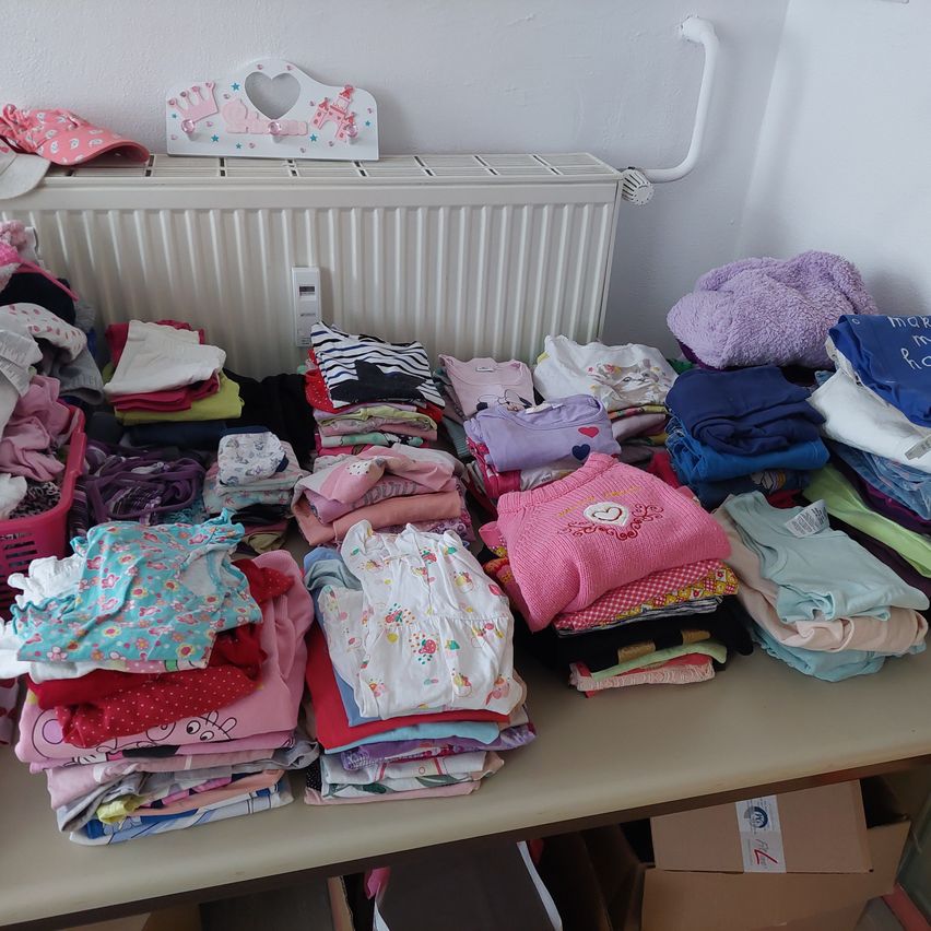 Ein überfüllter Raum mit einem Tisch voller gefalteter Babykleidung, einem weißen Heizkörper und verschiedenen Gegenständen wie einer rosa Mütze und einem weißen herzförmigen Objekt.
