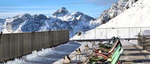 Eine hochgelegene Plattform mit Blick auf verschneite Berge, mit mehreren bunten Liegestühlen und einem Metallzaun. Der Himmel ist klar mit einigen Wolken.